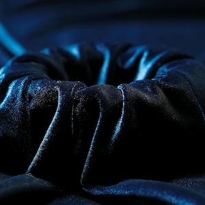 May include: Close-up of a dark blue scrunchie made of a soft, velvety fabric. The fabric is gathered and folded, creating a textured, circular shape. The lighting highlights the sheen of the material, emphasizing the deep color and the folds.