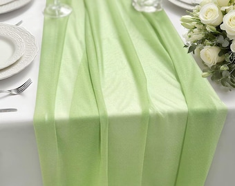 May include: A light green table runner made of a sheer, semi-transparent fabric with a smooth texture. The runner is draped vertically down the center of a white tablecloth, featuring soft folds and a consistent, solid pastel green color throughout.