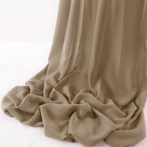 May include: A sheer, taupe-colored fabric drape featuring a lightweight, semi-transparent texture. The material hangs in soft, cascading folds that create natural vertical pleats and gentle curves as it pools toward the bottom of the frame.