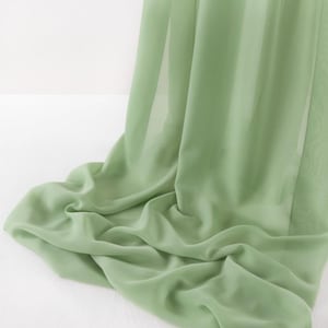May include: A sage green fabric with a sheer, lightweight, and semi-transparent texture. The material is draped in soft, flowing folds, creating a series of gentle curves and vertical lines that highlight the smooth, consistent color throughout.