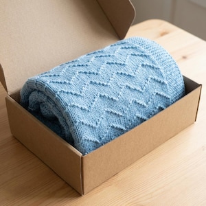 May include: A light blue knitted blanket with a chevron pattern is neatly folded inside a brown cardboard box. The blanket's texture and design are clearly visible, suggesting a cozy and handcrafted item. The box is open, revealing the blanket.