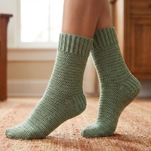 Könnte beinhalten: Ein Paar salbeigrüne Häkelsocken. Die Socken haben einen gerippten Bund und ein strukturiertes Maschenmuster. Die Socken werden an den Füßen einer Person gezeigt.