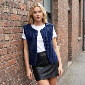May include: A navy blue knitted vest with an open front. The vest is worn over a white t-shirt and paired with a black leather skirt. The model is standing in front of a brick wall.