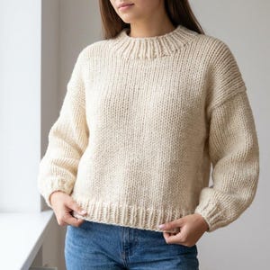 May include: Cream-colored, chunky knit sweater with a ribbed collar, cuffs, and hem. The sweater has a relaxed fit and long sleeves. The image shows a close-up of the sweater's texture and construction. The sweater is a cozy and warm garment.