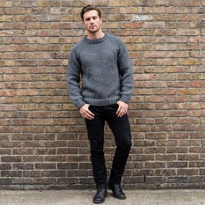 May include: A gray crew neck sweater and black jeans. The sweater has long sleeves and a ribbed hem. The jeans are a dark wash and have a slim fit. The person is standing in front of a brick wall.