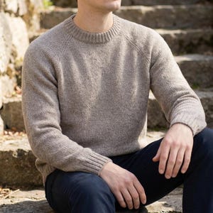 May include: A light brown, crew neck sweater with raglan sleeves. The sweater has a ribbed collar, cuffs, and hem. The person is wearing dark blue pants while sitting on stone steps.