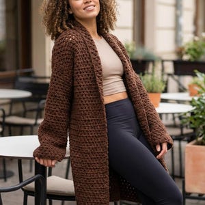 May include: A long, open-front brown crochet cardigan. The cardigan is made of thick yarn and has a textured appearance. The model is wearing a beige top and dark blue leggings. The background includes outdoor cafe tables and chairs.