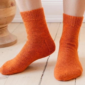 May include: A pair of orange knitted socks. The socks are a solid color and have a ribbed cuff. The socks are worn on a person's feet, with the feet resting on a light-colored wooden floor.
