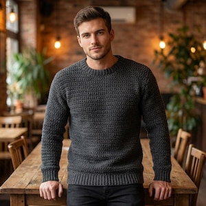 Könnte beinhalten: Ein dunkelgrauer Rundhals-Pullover mit einem strukturierten Strickmuster. Der Pullover hat gerippte Bündchen und einen Saum. Die Person steht in einem Restaurant und lehnt sich an einen Holztisch. Der Pullover ist einfarbig.