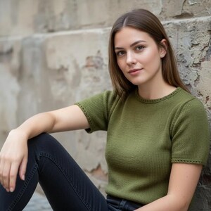 以下が含まれることがあります： オリーブグリーンの半袖ニットトップス。クルーネックとリブ編みのテクスチャが特徴です。濃い色のジーンズと合わせています。モデルは風化した壁に寄りかかっています。