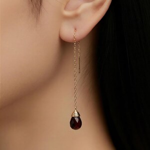 May include: A single gold-coloured earring featuring a long chain and a dark red, teardrop-shaped gemstone. The earring is suspended from the ear, highlighting the intricate design and the deep colour of the gem.