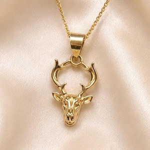Könnte beinhalten: Eine goldfarbene Halskette mit einem Anhänger in Form eines Hirschkopfes mit ausgeprägtem Geweih. Der Anhänger hängt an einer zarten goldfarbenen Kette. Das Metall hat eine polierte, reflektierende Oberfläche, die das Licht einfängt und die detaillierten Gesichtszüge sowie die geschwungene Struktur des Geweihs hervorhebt.