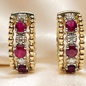 May include: A pair of hoop earrings crafted from yellow gold, featuring a central row of alternating round-cut red rubies and clear white diamonds. The edges of each hoop are detailed with a beaded texture, creating a distinct border.
