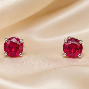 May include: A pair of round-cut, vibrant red gemstone stud earrings set in four-prong silver metal settings. Each earring features a faceted circular stone that reflects light, secured by a metallic base designed for pierced ears.