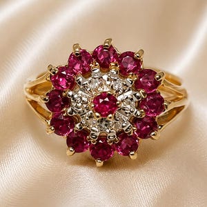 May include: A gold ring featuring a central cluster design with a round red gemstone surrounded by a halo of small white stones, encircled by a larger outer ring of round red gemstones, all set in a polished gold band with a split shank design.