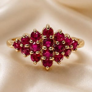 May include: A gold ring featuring a cluster of fifteen round, deep red rubies arranged in a marquise-shaped setting. The stones are held in place by small gold prongs, creating a dense, symmetrical floral-like pattern across the top of the band.