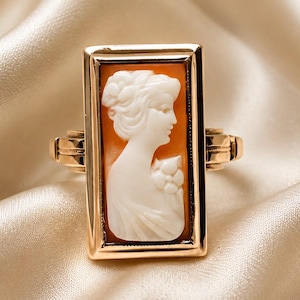 May include: A rectangular gold ring featuring a central cameo with a white profile portrait of a person against an orange background. The metal band has a polished finish and a structured frame that holds the carved cameo securely in place.