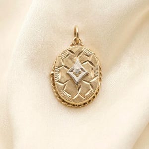 May include: An oval gold locket pendant featuring a textured, cross-hatched surface pattern and a central white gold diamond-shaped inlay. A single small clear stone is set in the centre of the inlay, and the locket has a twisted rope border.