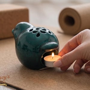 Puede incluir: Un difusor de aceites esenciales de cerámica en forma de pájaro verde azulado con una vela de té encendida en su interior. El pájaro tiene agujeros decorativos en la parte superior. Una mano coloca la vela en el difusor. Una esterilla y un bloque de yoga están en el fondo.