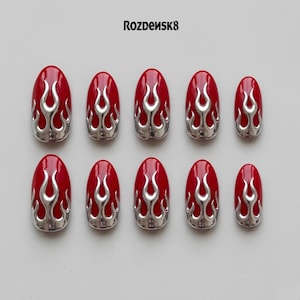 May include: A set of ten oval-shaped press-on nails featuring a glossy red base with a raised, metallic silver flame design. The text Rozdensk8 is printed in black at the top center. The nails are arranged in two rows of five on a plain background.