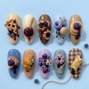 May include: Ten artificial nail tips featuring 3D miniature food designs including blueberry pie slices, a spoon with jam, pastries, macarons, pancakes, a croissant, cubed fruit, a donut with a whisk, and a cannoli on a brown checkered base.