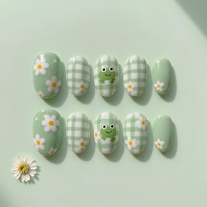 May include: A set of ten oval press-on nails in a light sage green base color. The designs include white daisy flowers with yellow centers, a light green and white gingham check pattern, and a small green frog face with black eyes and a smile.