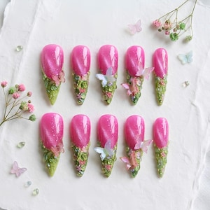 May include: A set of ten press-on nails featuring a vibrant pink cat-eye finish on the upper half and a textured green base adorned with small, translucent butterfly embellishments and tiny green stone accents arranged in a pointed shape.