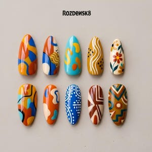 May include: Ten almond-shaped press-on nails featuring diverse designs including abstract shapes, wavy lines, floral patterns, and geometric motifs in blue, orange, yellow, brown, and cream. The text ROZDENSK8 is printed at the top center.