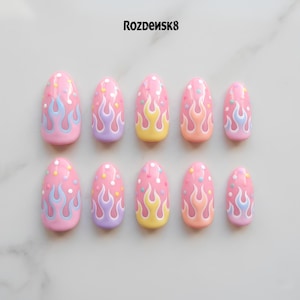 May include: A set of ten pink press-on nails with a flame design. Each nail features a pink base with colorful flames in blue, yellow, and purple. The nails are oval-shaped and have a glossy finish. The text "Rozdeusk8" is at the top.