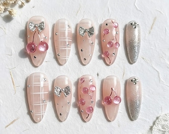 May include: A set of ten press-on nails featuring a light pink base with various designs including white plaid patterns, silver glitter accents, small pink cherry charms, and silver bow embellishments, all accented with tiny clear rhinestones.