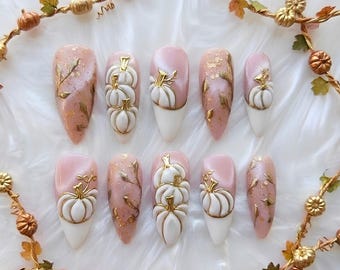 May include: A set of ten press-on nails featuring a soft pink and white color palette. The designs include raised white pumpkin motifs with gold stems, gold leaf accents, and delicate gold vine detailing across the almond-shaped nail tips.