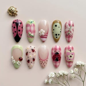 May include: Ten almond-shaped press-on nails featuring various designs including ladybugs, pink gingham patterns, green apples, white flowers, and a caterpillar. A small gold ladybug charm is positioned separately above the nail set.