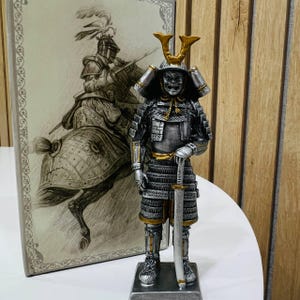 May include: A silver and gold samurai figurine standing on a rectangular base, wearing detailed armour and a helmet with a prominent crest, holding a katana. Behind the figurine is a rectangular panel featuring a sketch of a mounted knight.