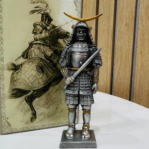 May include: A standing samurai figurine in silver and black armour with gold accents, featuring a crescent moon crest on the helmet and a sword held in the right hand. The figure stands on a rectangular base, positioned before a decorative panel.