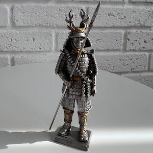 May include: A silver-coloured samurai figurine standing on a rectangular base. The warrior wears detailed plate armour with gold accents and a helmet featuring prominent antler-like crests. A long spear is held upright in the right hand.