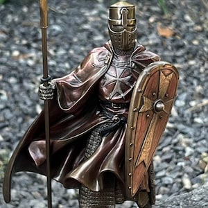 May include: A bronze-toned knight figurine standing on a textured base. The knight wears a full helmet, chainmail, and a flowing cloak with a cross emblem on the chest. One hand holds a tall spear, while the other grips a wooden-textured shield.