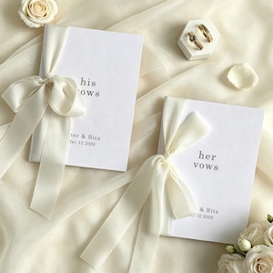 Può includere: Due libretti di promesse bianchi legati con nastri color avorio, uno con la scritta "his vows" e l'altro "her vows". Fedi nuziali dorate poggiano su un supporto esagonale bianco. I libri sono stampati con nomi e una data. Accenti floreali completano la scena.