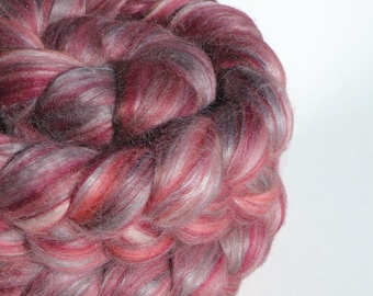 May include: A close-up shot of a circular bundle of hand-dyed yarn. The yarn is a mix of burgundy, pink, and grey fibres, creating a textured, multi-tonal appearance. The yarn is tightly wound, showcasing the colour variations.