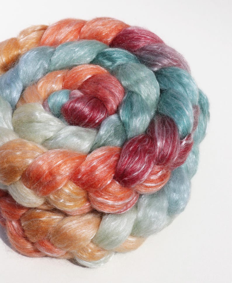 May include: A close-up of a circular braid of colorful yarn. The yarn is a mix of colors, including orange, red, teal, and blue. The braid is tightly woven and has a soft, textured appearance. The yarn is likely used for knitting or crocheting.