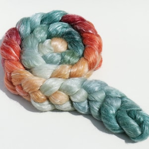 May include: A braided skein of colorful yarn in a spiral shape. The yarn transitions from red to orange, then to peach, light green, and finally, a deep teal. The yarn is soft and has a slight sheen, suggesting a luxurious fiber.