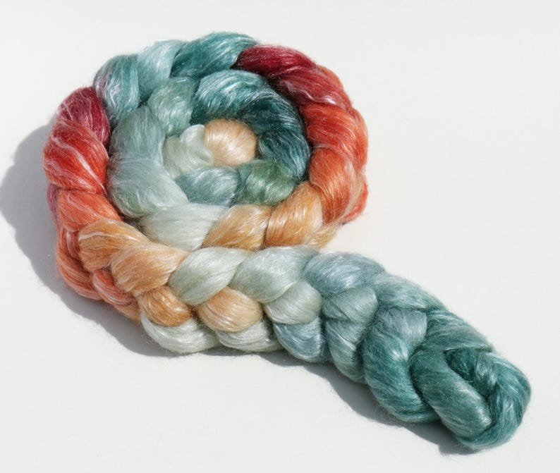 May include: A braided skein of colorful yarn in a spiral shape. The yarn transitions from red to orange, then to peach, light green, and finally, a deep teal. The yarn is soft and has a slight sheen, suggesting a luxurious fiber.