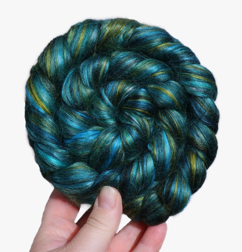 Peut inclure: Une main tenant une pelote de fibres turquoise, vertes et dor&eacute;es. La fibre est fil&eacute;e en un fil &eacute;pais et moelleux.