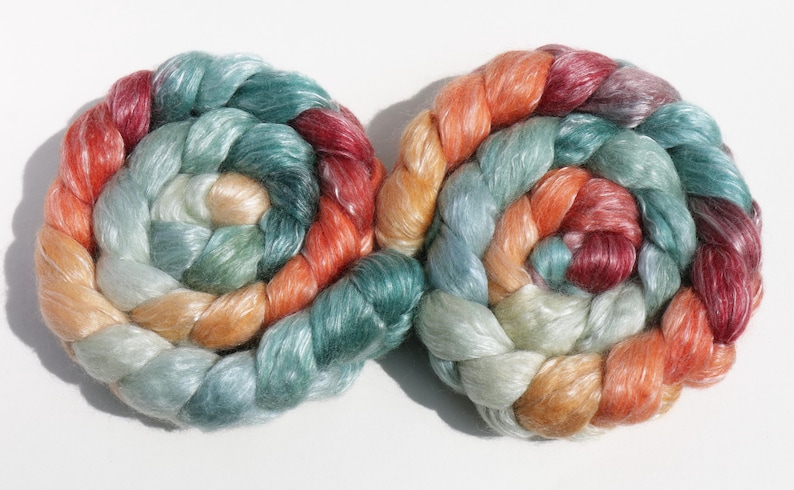 May include: Two spiral-shaped skeins of colorful yarn. The yarn is a blend of colors including red, orange, teal, and cream. The yarn is tightly wound, creating a textured appearance. The skeins are displayed on a white background.