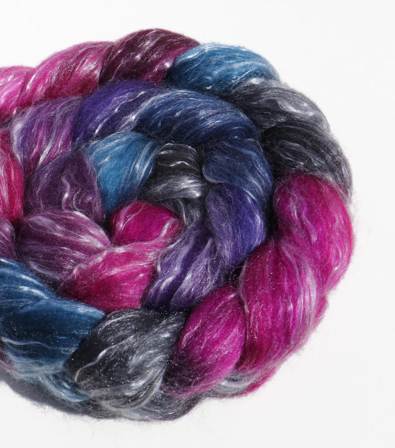 K&ouml;nnte beinhalten: Ein dicker, geflochtener Strang aus Fasern mit einer bunten Farbpalette aus tiefem Magenta, K&ouml;nigsblau, dunklem Anthrazit und hellem Violett. Die Fasern haben einen deutlichen Glanz und eine weiche, schimmernde Textur, kreisf&ouml;rmig gewickelt.