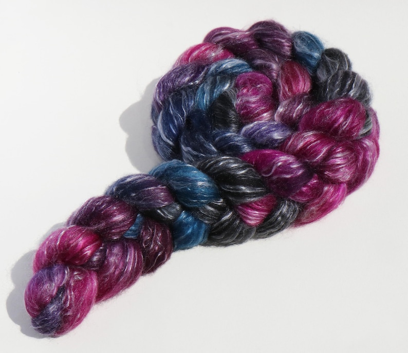 K&ouml;nnte beinhalten: Ein Strang handgef&auml;rbtes Garn mit einer bunten Farbpalette aus tiefem Magenta, K&ouml;nigsblau, Anthrazit und Schwarz. Die Faser hat einen ausgepr&auml;gten Glanz und ist zu einer dicken, geflochtenen Struktur mit sichtbarer Fasertextur verdreht.