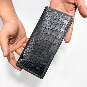 Può includere: Un portafoglio rettangolare nero caratterizzato da una texture a pelle di coccodrillo su tutta la superficie. Il portafoglio presenta cuciture visibili lungo i bordi e una forma sottile e allungata con una finitura goffrata molto dettagliata.