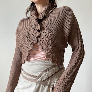 Puede incluir: Un bolero de punto marrón con mangas largas y cuello de ganchillo festoneado, usado sobre una blusa rosa pálido y una falda cruzada beige claro con bandas de tela diagonales y un pequeño detalle metálico circular en la cintura.