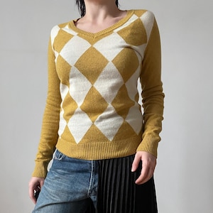 Peut inclure: Un pull à manches longues et col en V avec un motif losange argyle jaune moutarde et blanc, associé à un pantalon hybride combinant un jean en denim bleu d'un côté et un panneau en tissu noir plissé de l'autre côté.
