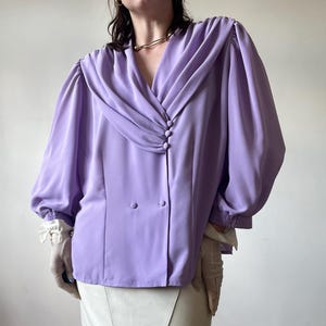 Puede incluir: Una blusa de color lila con cuello drapeado y mangas largas abullonadas. La blusa presenta un diseño cruzado con tres botones y se combina con una falda color crema. La blusa tiene una estética vintage.