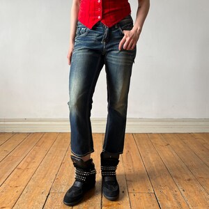 May include: A bright red sleeveless vest featuring a V-neckline and a vertical pinstripe pattern, paired with dark blue denim jeans that have a faded wash and straight-leg cut, along with black high-top sneakers adorned with silver metal studs.